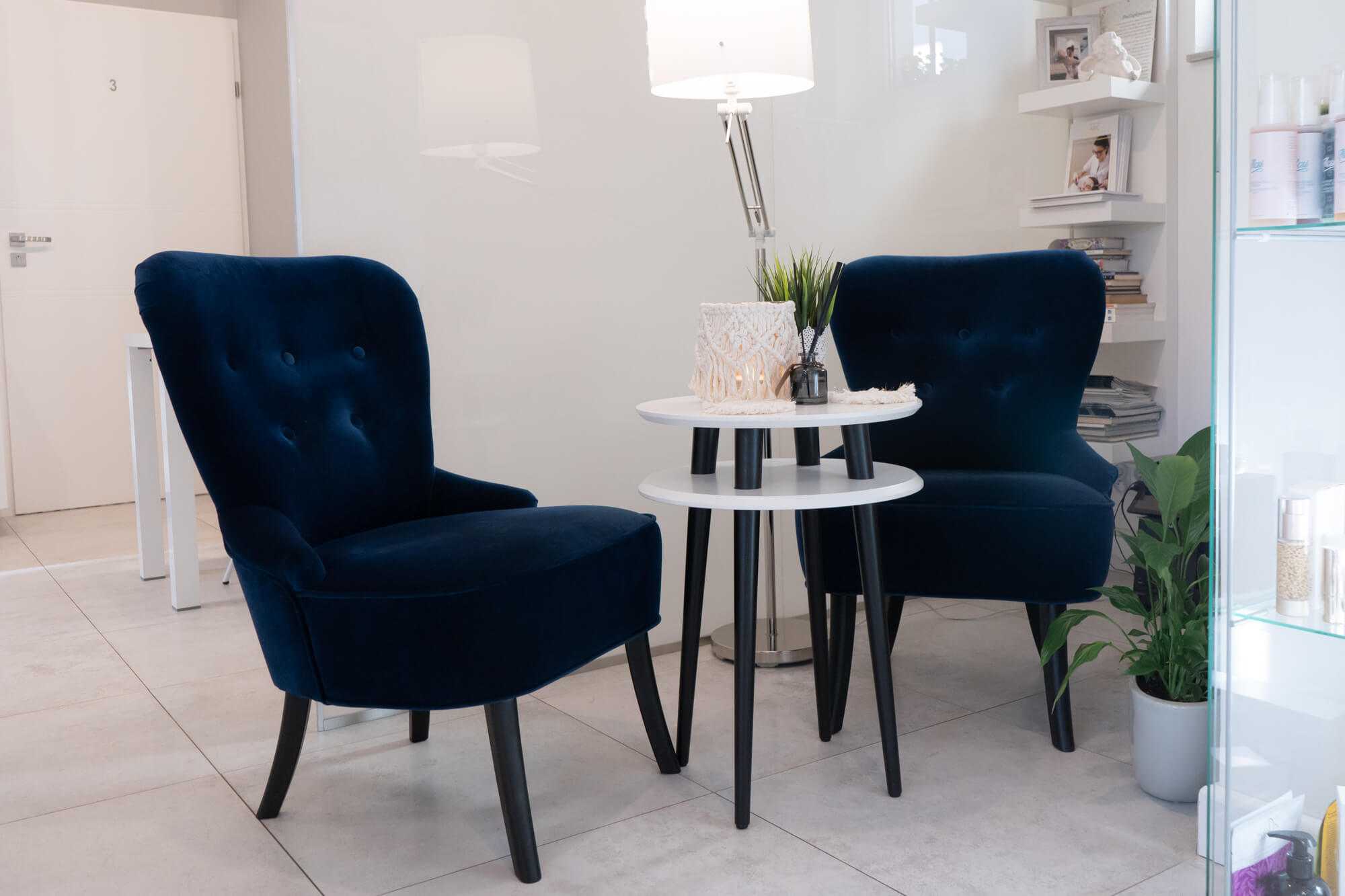 Poczekalnia w Salonie Kosmetycznym „Evita” z granatowymi fotelami, stolikiem kawowym, kwiatami i półką z magazynami
